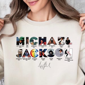 May include: Cream-colored sweatshirt featuring the name "MICHAEL JACKSON" in a colorful, collage-style design. The design includes images and dates from his career, along with a signature. The sweatshirt is worn by a person.