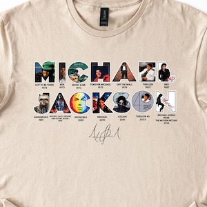 May include: A beige t-shirt featuring the name "MICHAEL JACKSON" in a colorful, collage-style design. Each letter is composed of images from his album covers and music videos. Below the name is a signature. The shirt is made of a soft material.