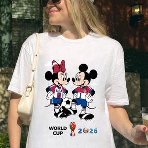 May include: White t-shirt featuring Mickey and Minnie Mouse in soccer uniforms, with a soccer ball between them. The shirt has the text "WORLD CUP 2026" in blue and red. The shirt is worn by a person.