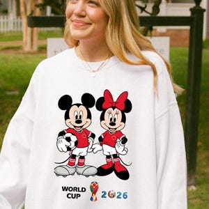 May include: White sweatshirt featuring Mickey and Minnie Mouse in soccer uniforms, holding a soccer ball. The design includes the text "WORLD CUP 2026" in blue and yellow. The characters are wearing red shirts and shorts.