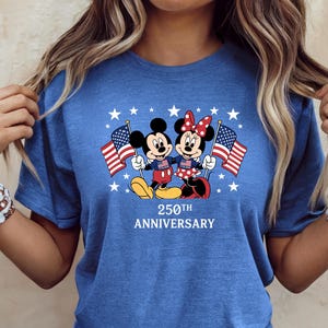 Pu&ograve; includere: Una t-shirt blu a maniche corte con una grafica di Topolino e Minnie che tengono in mano bandiere americane. Il design include stelle bianche e il testo 250TH ANNIVERSARY stampato in bianco sotto i personaggi. Girocollo.