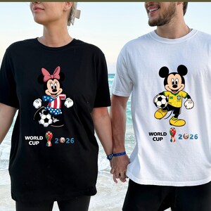 Puede incluir: Camisetas negras y blancas con dise&ntilde;os de Mickey y Minnie Mouse sosteniendo balones de f&uacute;tbol. Minnie Mouse lleva un lazo rosa y los colores de la bandera estadounidense. Mickey Mouse luce un dise&ntilde;o con los colores de la bandera brasile&ntilde;a. Ambas camisetas muestran "WORLD CUP 2026".