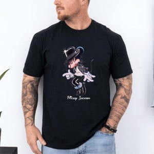 May include: A black short-sleeved t-shirt featuring a graphic of a cartoon mouse dressed in a black fedora, white shirt, and black jacket, performing a dance move. The text "Mickey Jackson" is printed in white below the graphic illustration.