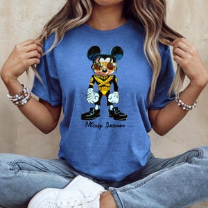 Puede incluir: Una camiseta azul de manga corta con un gr&aacute;fico de un rat&oacute;n de dibujos animados vestido con una chaqueta militar negra y dorada con gafas de sol amarillas. El gr&aacute;fico incluye el texto Mickey Jackson escrito en cursiva negra debajo.
