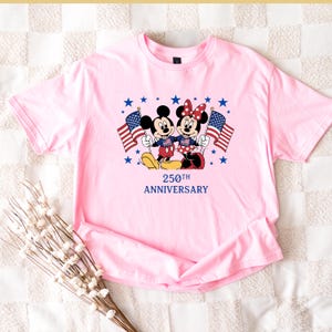 Pu&ograve; includere: Una t-shirt rosa chiaro con una grafica di Topolino e Minnie che tengono in mano bandiere americane. I personaggi sono circondati da stelle blu e il testo 250TH ANNIVERSARY &egrave; stampato in blu. La maglietta presenta un girocollo.
