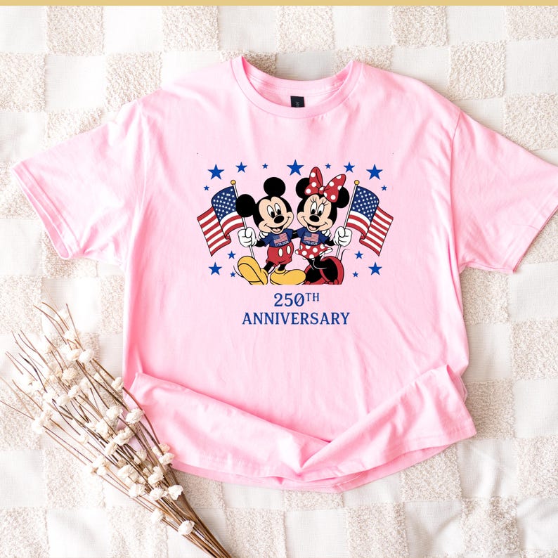 Pu&ograve; includere: Una t-shirt rosa chiaro con una grafica di Topolino e Minnie che tengono in mano bandiere americane. I personaggi sono circondati da stelle blu e il testo 250TH ANNIVERSARY &egrave; stampato in blu. La maglietta presenta un girocollo.