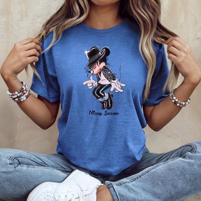 May include: A blue short-sleeve t-shirt featuring a graphic of a cartoon mouse dressed in a black fedora, black jacket, and black trousers, performing a dance move. The text "Mickey Jackson" is printed in black lettering below the graphic.
