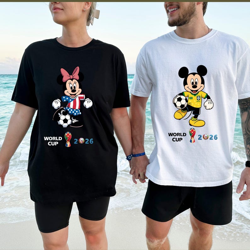 Puede incluir: Camisetas negras y blancas con dise&ntilde;os de Mickey y Minnie Mouse sosteniendo balones de f&uacute;tbol. Minnie Mouse lleva un lazo rosa y los colores de la bandera estadounidense. Mickey Mouse luce un dise&ntilde;o con los colores de la bandera brasile&ntilde;a. Ambas camisetas muestran "WORLD CUP 2026".