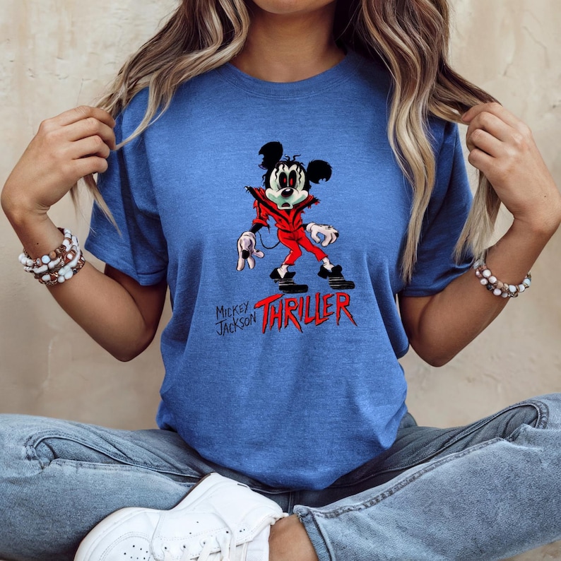 May include: A blue short-sleeve t-shirt featuring a graphic of a cartoon mouse dressed in a red jacket. The character has dark hair and white gloves. Below the graphic, the text reads Mickey Jackson THRILLER in black and red lettering.