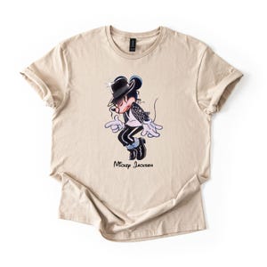 May include: A beige short-sleeved t-shirt featuring a graphic of a cartoon mouse dressed in a black fedora, white shirt, and black jacket, performing a dance move. Below the character, the text Mickey Jackson is printed in black script.