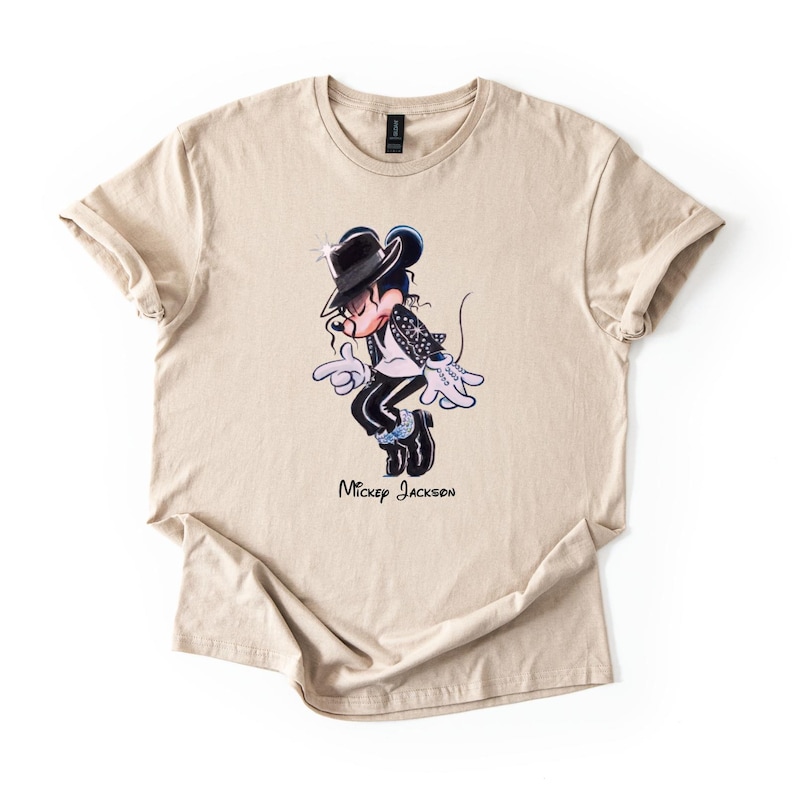 May include: A beige short-sleeved t-shirt featuring a graphic of a cartoon mouse dressed in a black fedora, white shirt, and black jacket, performing a dance move. Below the character, the text Mickey Jackson is printed in black script.