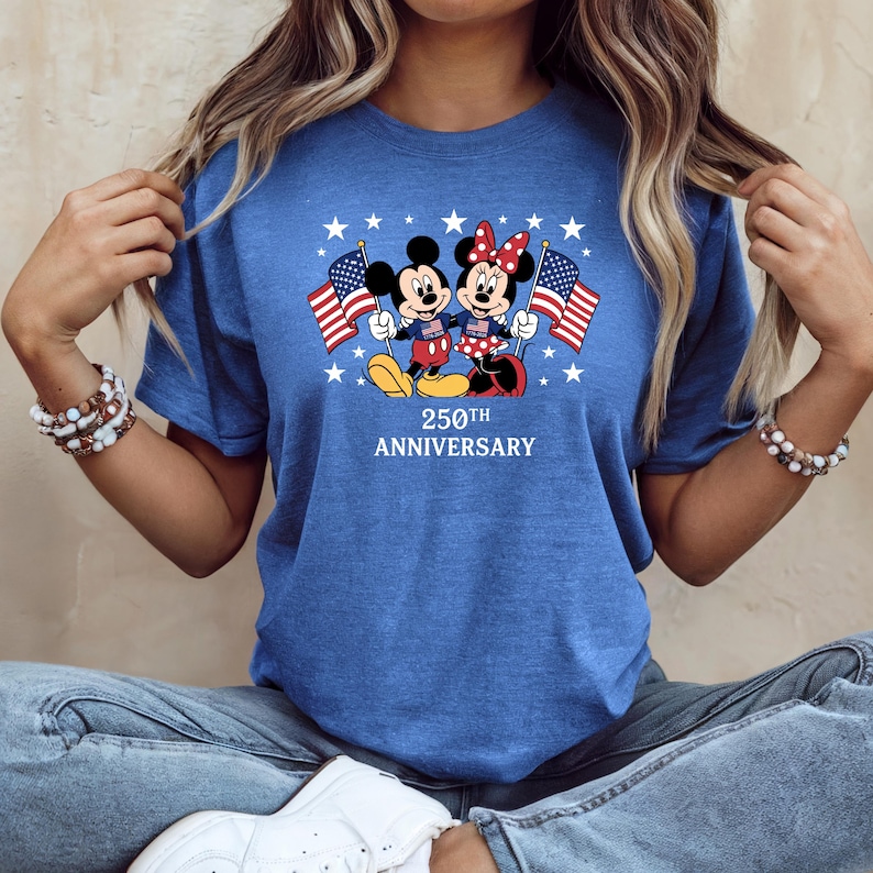 Pu&ograve; includere: Una t-shirt blu a maniche corte con una grafica di Topolino e Minnie che tengono in mano bandiere americane. Il design include stelle bianche e il testo 250TH ANNIVERSARY stampato in bianco sotto i personaggi. Girocollo.
