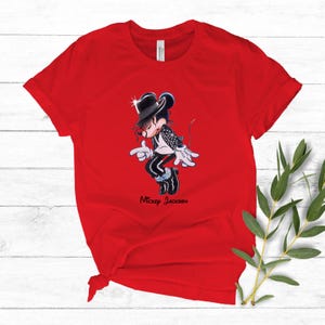 May include: A bright red short-sleeved t-shirt featuring a graphic of a cartoon mouse dressed in a black fedora, white shirt, and black jacket with white accents. The character is in a dancing pose with the text "Mickey Jackson" printed below.