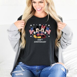 Pu&ograve; includere: Una t-shirt nera con una grafica di Topolino e Minnie che tengono bandiere americane, circondati da stelle bianche. Sotto i personaggi, il testo 250TH ANNIVERSARY &egrave; scritto in lettere maiuscole bianche. Indossata con un cardigan grigio.