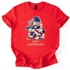 May include: A red t-shirt featuring Mickey Mouse, Donald Duck, and Goofy holding a flag with the American flag design. The text "250th Anniversary" is printed below the characters. The shirt is made of a soft material.