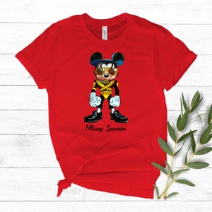 Puede incluir: Una camiseta roja de manga corta con el gr&aacute;fico de un rat&oacute;n de dibujos animados vestido con una chaqueta militar negra y dorada con gafas de sol amarillas. Debajo del personaje, el texto Mickey Jackson est&aacute; impreso en letra cursiva negra.