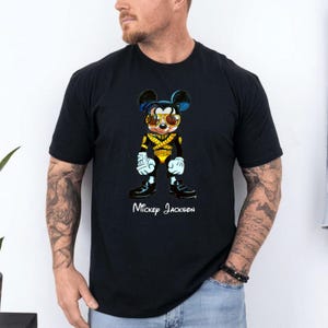 Puede incluir: Una camiseta negra de manga corta con el gr&aacute;fico de un rat&oacute;n de dibujos animados vestido con una chaqueta de estilo militar negra y dorada y gafas de sol amarillas. Debajo del personaje, el texto blanco dice Mickey Jackson en cursiva.