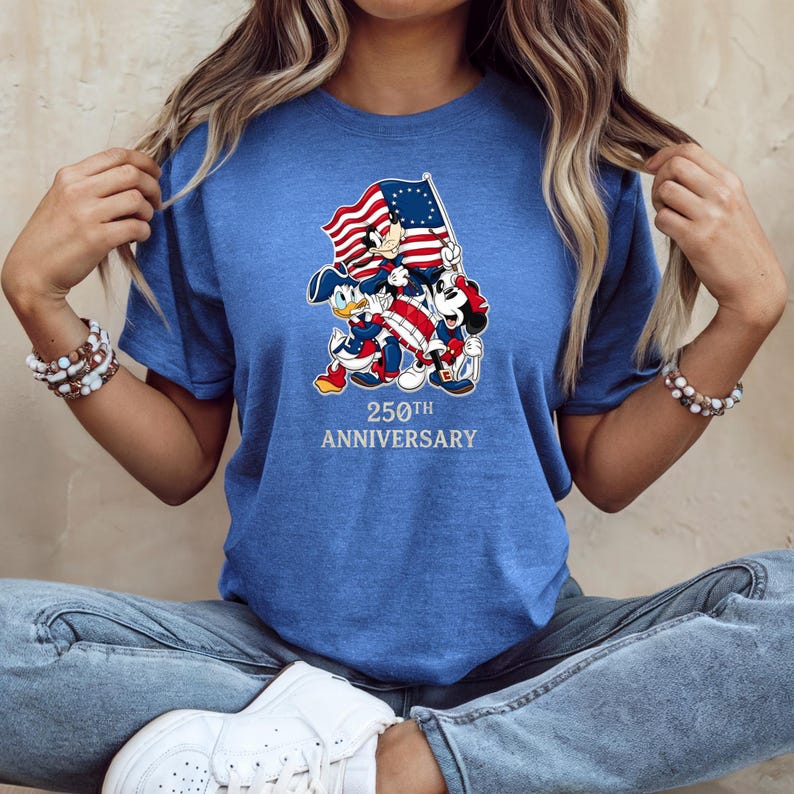 May include: Blue t-shirt featuring a graphic of Mickey Mouse, Donald Duck, and Goofy holding an American flag. The graphic also includes the text "250th Anniversary" in silver lettering.