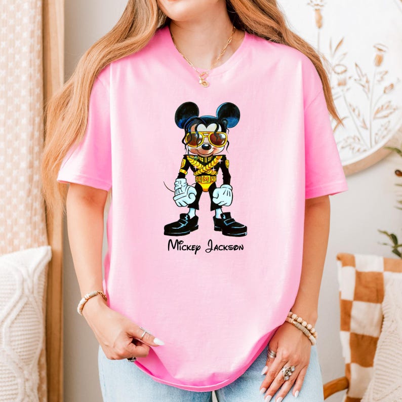 Puede incluir: Una camiseta de manga corta de color rosa claro con un gr&aacute;fico de un rat&oacute;n de dibujos animados vestido con una chaqueta militar negra y dorada y gafas de sol. El texto "Mickey Jackson" est&aacute; impreso en letras cursivas negras bajo el gr&aacute;fico.