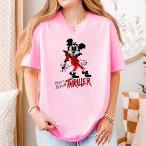 May include: A light pink short-sleeved t-shirt featuring a graphic of a cartoon mouse dressed in a red jacket with the text Mickey Jackson THRILLER printed in black and red lettering below the character on the front of the garment.