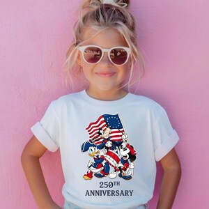 May include: A white t-shirt featuring a graphic of Mickey Mouse, Donald Duck, and Goofy holding a flag with red and white stripes and blue stars. The text "250th Anniversary" is below the graphic.