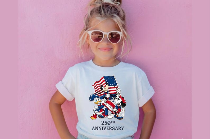 May include: A white t-shirt featuring a graphic of Mickey Mouse, Donald Duck, and Goofy holding a flag with red and white stripes and blue stars. The text "250th Anniversary" is below the graphic.