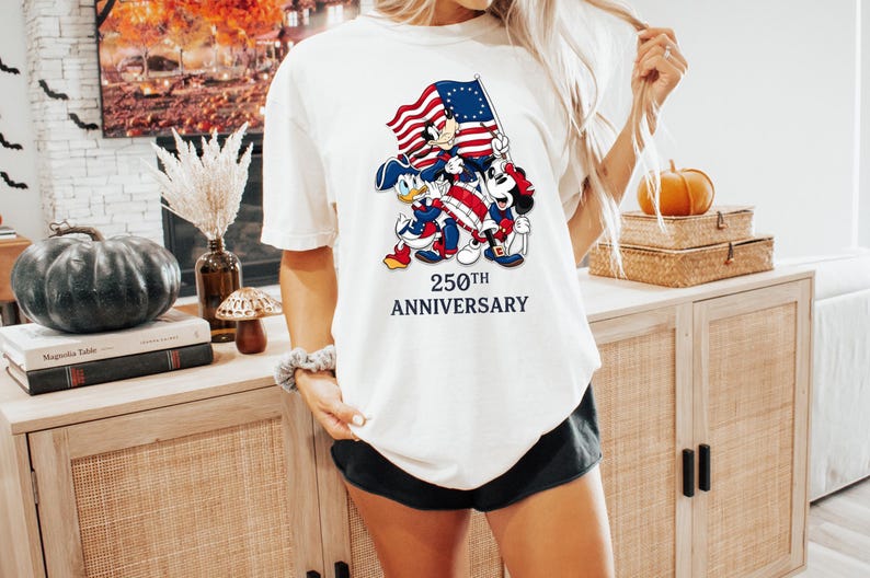 May include: Cream-colored t-shirt featuring a graphic of Mickey Mouse, Donald Duck, and Minnie Mouse holding an American flag. The text "250th ANNIVERSARY" is below the characters. The shirt is paired with black shorts.