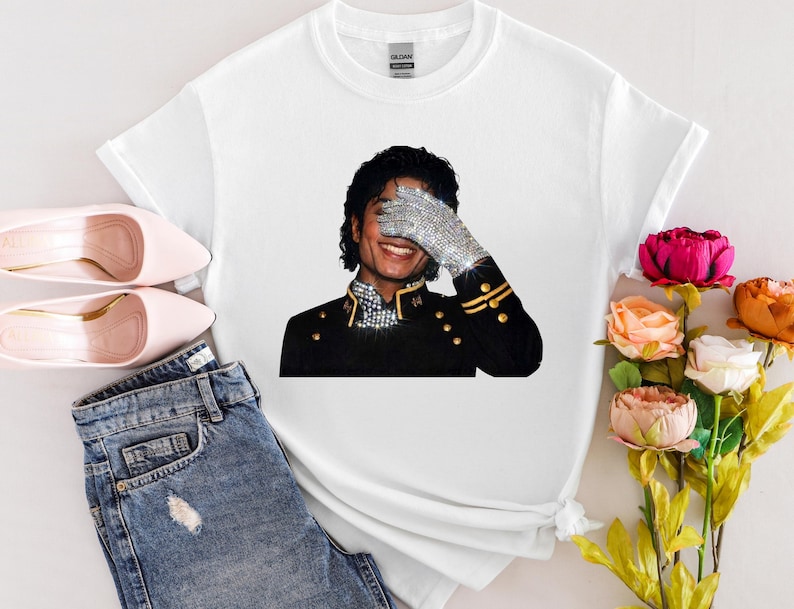 May include: White t-shirt with a graphic of Michael Jackson, wearing a sparkling glove and black jacket. The image is set against a neutral background with pink shoes and denim jeans.