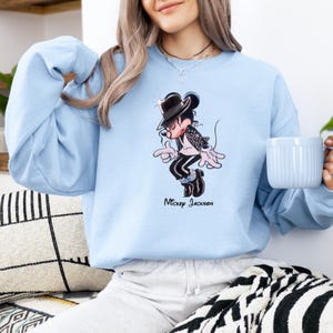 May include: A light blue crewneck sweatshirt featuring a graphic illustration of a cartoon mouse dressed in a black fedora, sequined jacket, and black trousers, performing a dance move. The text "Mickey Jackson" is printed below the graphic.