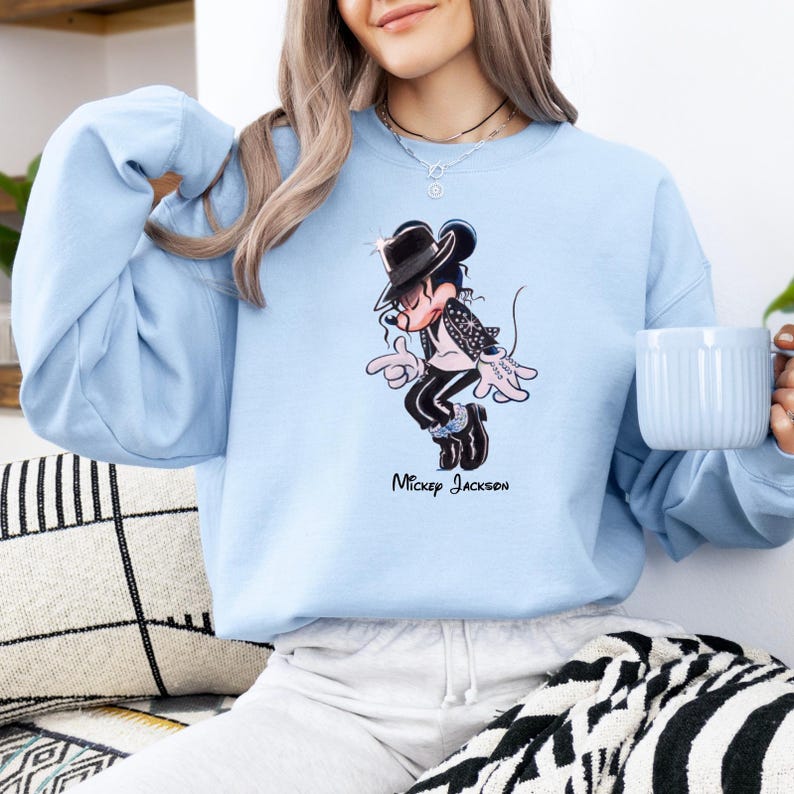 May include: A light blue crewneck sweatshirt featuring a graphic illustration of a cartoon mouse dressed in a black fedora, sequined jacket, and black trousers, performing a dance move. The text "Mickey Jackson" is printed below the graphic.