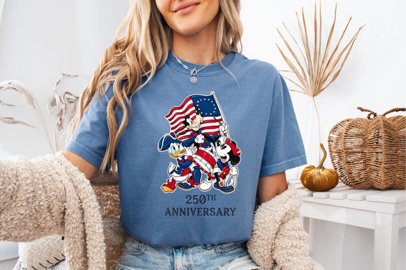 May include: A blue t-shirt featuring a graphic of cartoon characters holding an American flag. The graphic includes Mickey Mouse, Minnie Mouse, and Donald Duck. The text "250th ANNIVERSARY" is below the graphic.