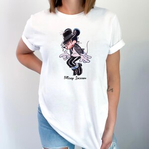 May include: A white short-sleeved t-shirt featuring a graphic illustration of a cartoon mouse dressed in a black fedora, black jacket, and black trousers, performing a dance move. The text "Mickey Jackson" is printed in black below the illustration.