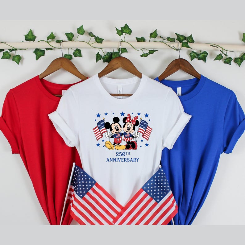 Pu&ograve; includere: Una t-shirt bianca con una grafica di Topolino e Minnie che tengono bandiere americane, con il testo 250TH ANNIVERSARY stampato sotto. La maglietta &egrave; esposta tra una t-shirt rossa tinta unita e una blu tinta unita su grucce di legno.