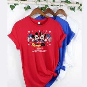Pu&ograve; includere: Una t-shirt rossa a maniche corte con una grafica di Topolino e Minnie che tengono bandiere americane circondati da stelle bianche. Sotto i personaggi, il testo recita 250TH ANNIVERSARY. La maglietta &egrave; appesa a una gruccia di legno.
