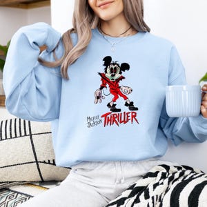 May include: A light blue crewneck sweatshirt featuring a graphic of a cartoon mouse dressed in a red jacket, black trousers, and black shoes. The text "MICKEY JACKSON" appears in black, with the word "THRILLER" printed in large, bold red letters below.
