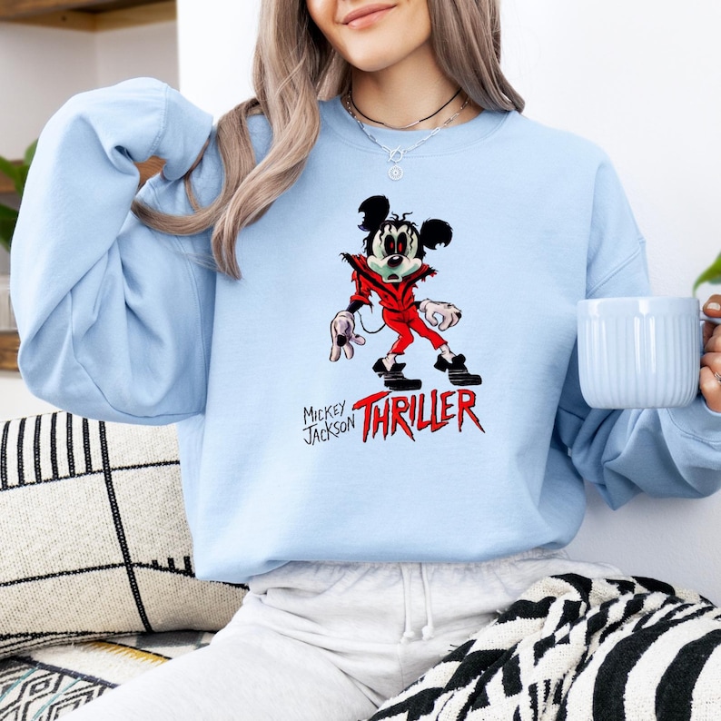 May include: A light blue crewneck sweatshirt featuring a graphic of a cartoon mouse dressed in a red jacket, black trousers, and black shoes. The text "MICKEY JACKSON" appears in black, with the word "THRILLER" printed in large, bold red letters below.