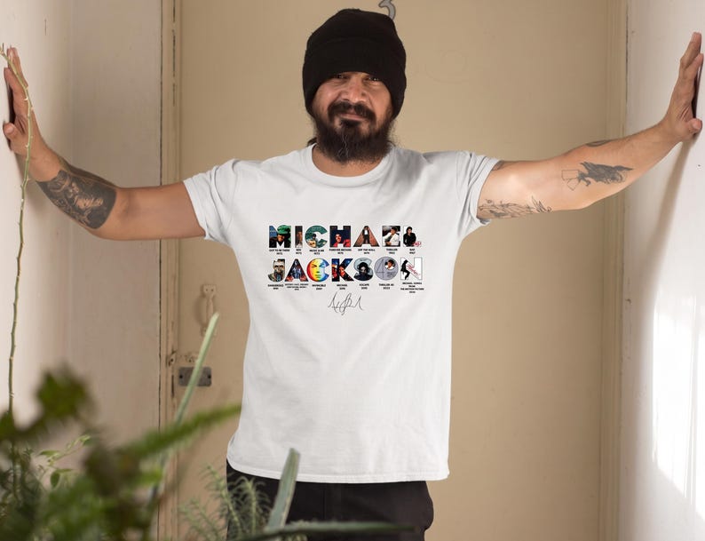 May include: A white short-sleeved t-shirt featuring a graphic print of the text MICHAEL JACKSON. The letters contain small images of album covers and music videos, with a signature printed below the text in the center of the chest area.