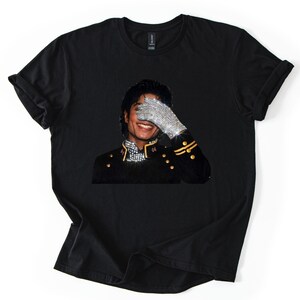 May include: Black t-shirt with a graphic of a smiling person wearing a bejeweled glove, covering their eyes. The person is wearing a black military-style jacket with gold detailing. The image is centered on the shirt.