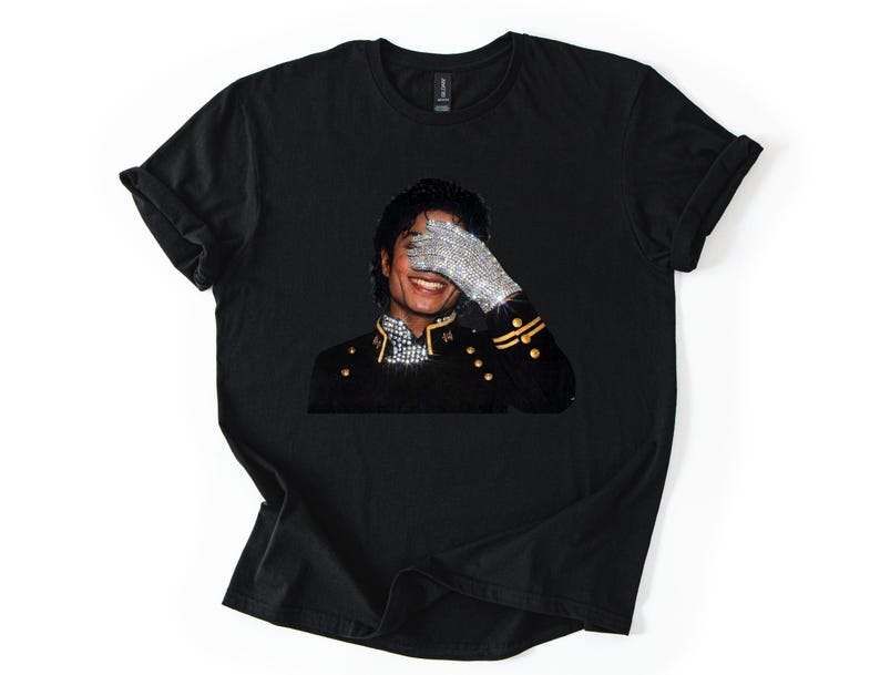 May include: Black t-shirt with a graphic of a smiling person wearing a bejeweled glove, covering their eyes. The person is wearing a black military-style jacket with gold detailing. The image is centered on the shirt.