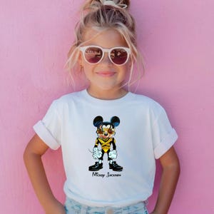 Puede incluir: Una camiseta blanca de manga corta con un estampado gr&aacute;fico de un rat&oacute;n de dibujos animados vestido con una chaqueta de estilo militar negra y dorada con gafas de sol. El texto Mickey Jackson est&aacute; impreso en negro bajo la ilustraci&oacute;n.