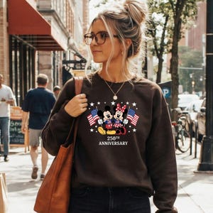 Pu&ograve; includere: Una felpa a maniche lunghe marrone scuro con una grafica di Topolino e Minnie che tengono bandiere americane, circondati da stelle bianche. Sotto i personaggi, il testo recita 250TH ANNIVERSARY. La felpa ha un girocollo.