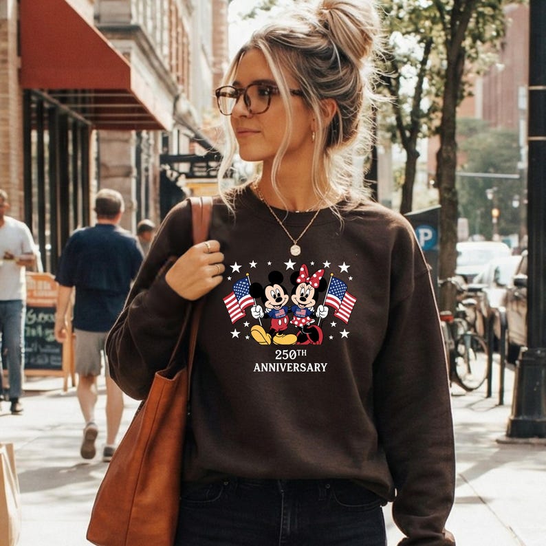 Pu&ograve; includere: Una felpa a maniche lunghe marrone scuro con una grafica di Topolino e Minnie che tengono bandiere americane, circondati da stelle bianche. Sotto i personaggi, il testo recita 250TH ANNIVERSARY. La felpa ha un girocollo.