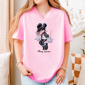 May include: A light pink short-sleeved t-shirt featuring a graphic of a cartoon mouse dressed in a black fedora, a black jacket with silver accents, and black trousers, performing a dance move. The text "Mickey Jackson" is printed below the graphic.
