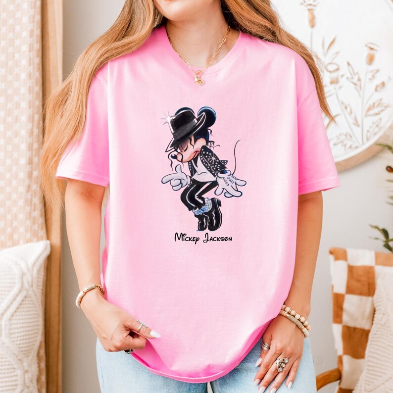 May include: A light pink short-sleeved t-shirt featuring a graphic of a cartoon mouse dressed in a black fedora, a black jacket with silver accents, and black trousers, performing a dance move. The text "Mickey Jackson" is printed below the graphic.