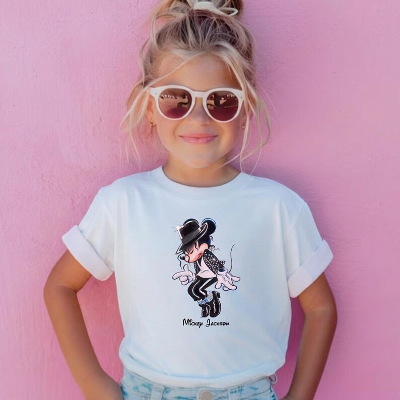 May include: A white short-sleeved t-shirt featuring a graphic print of a cartoon mouse dressed in a black fedora, black jacket, and black trousers with white stripes. The graphic includes the text "Mickey Jackson" in a black script font.