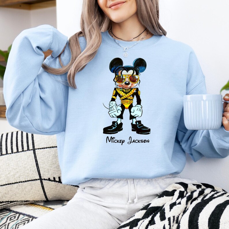 Puede incluir: Una sudadera de cuello redondo azul claro con un gr&aacute;fico de un rat&oacute;n de dibujos animados vestido con una chaqueta militar negra y dorada con gafas de sol. El personaje aparece en una pose con el texto Mickey Jackson impreso en negro.