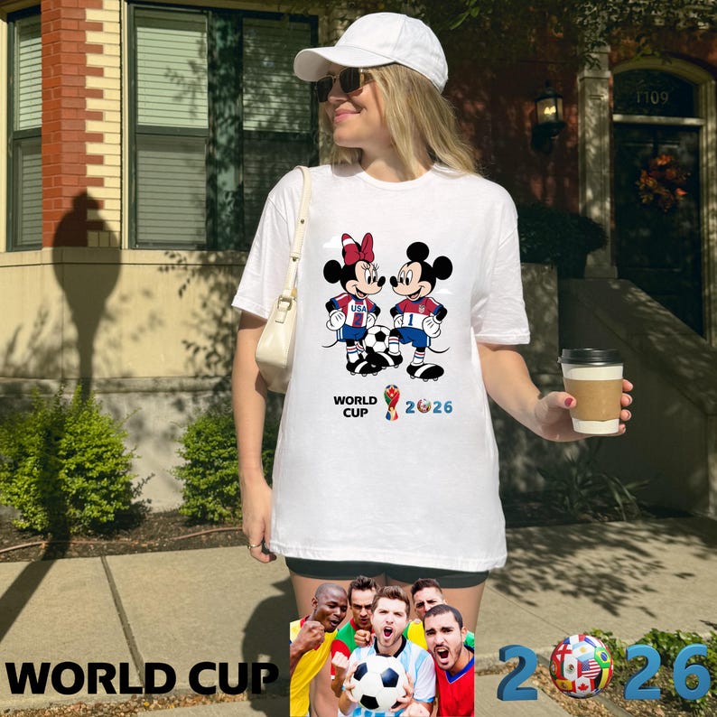 May include: White t-shirt featuring Mickey and Minnie Mouse in soccer uniforms, with a soccer ball between them. The shirt has the text "WORLD CUP 2026" in blue and red. The shirt is worn by a person.