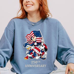 May include: A light blue sweatshirt featuring Mickey Mouse, Donald Duck, and Minnie Mouse holding a flag with red and white stripes and a blue field of stars. The text "250th Anniversary" is printed below the characters.