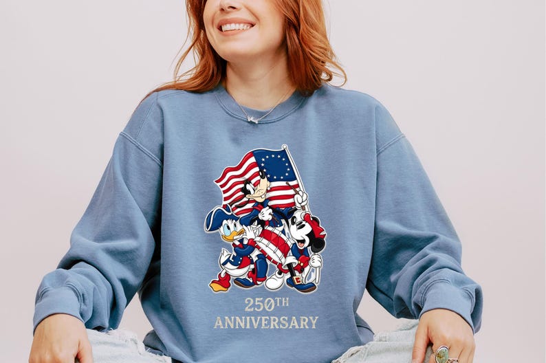 May include: A light blue sweatshirt featuring Mickey Mouse, Donald Duck, and Minnie Mouse holding a flag with red and white stripes and a blue field of stars. The text "250th Anniversary" is printed below the characters.