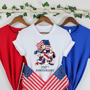 May include: Three t-shirts in red, white, and blue hang on a wooden rod. The white t-shirt features a graphic of Mickey Mouse, Donald Duck, and Goofy holding a flag with the text "250th Anniversary." Two American flags are in the foreground.